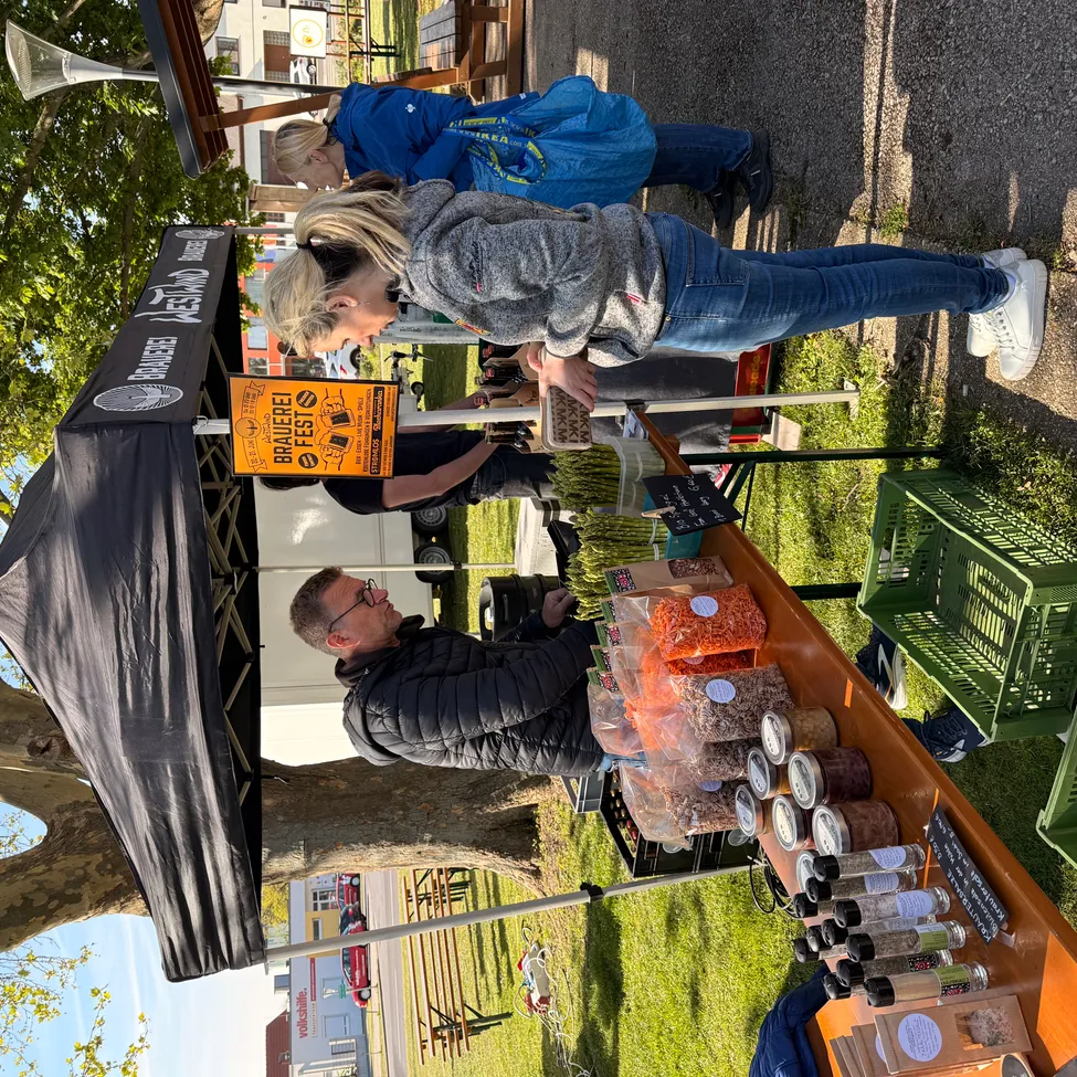 Ein Lebensmittelstand mit schwarzem Baldachin. Ein Mann und eine Frau schauen sich das Essen an. Ein Schild mit dem Text 'BRAUEPEL FEST' hängt am Stand.