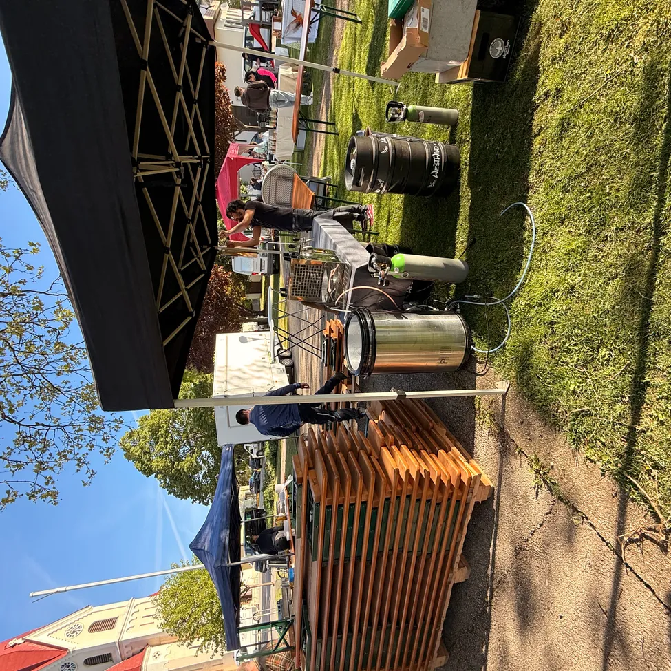 Ein Food-Stand auf einem Rasen mit schwarzem Zelt, Fässern und Tischen. Menschen sind beim Arbeiten unter dem Zelt zu sehen. Bäume und ein Gebäude sind im Hintergrund sichtbar.