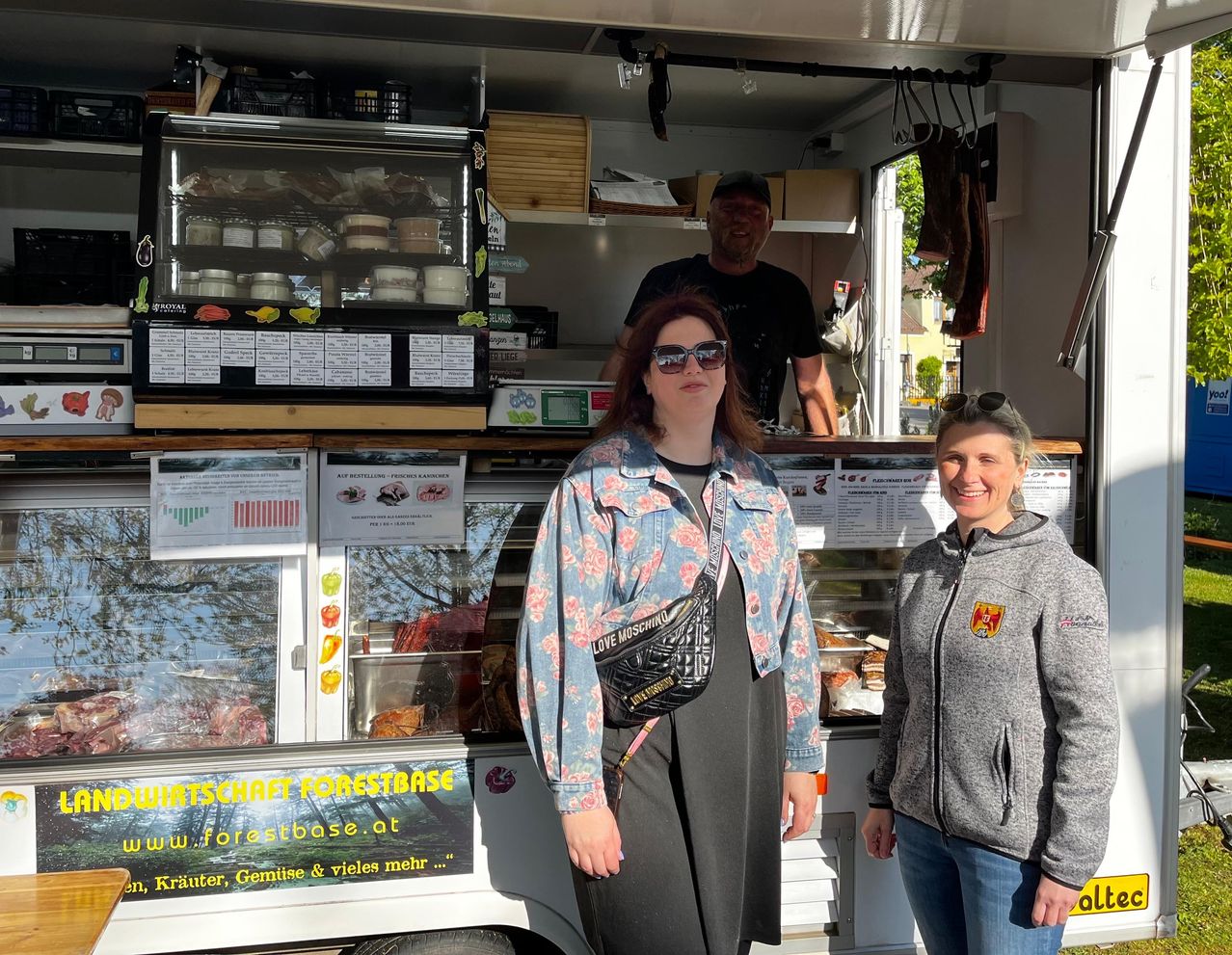 Drei Menschen stehen vor einem Food-Truck mit einem Menübrett, das 'Forestbase' beschriftet ist. Eine Frau in einer Blumenjacke und ein Mann in einem grauen Sweatshirt lächeln für die Kamera.