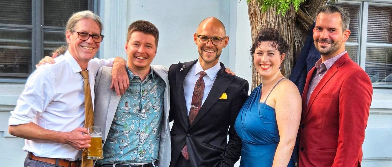 Fünf Personen, drei Männer und zwei Frauen, stehen eng beieinander und lächeln. Der Mann auf der linken Seite hält ein Glas Bier. Der Mann in der Mitte trägt einen Anzug und die Frau auf der rechten Seite trägt ein blaues Kleid.