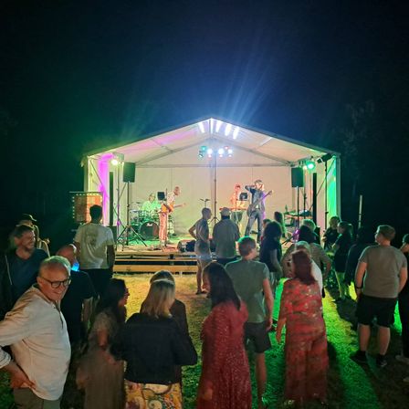 Eine Band spielt auf einer Bühne unter einem Zelt mit bunten Lichtern, während eine Menge Menschen bei Nacht zuschaut.