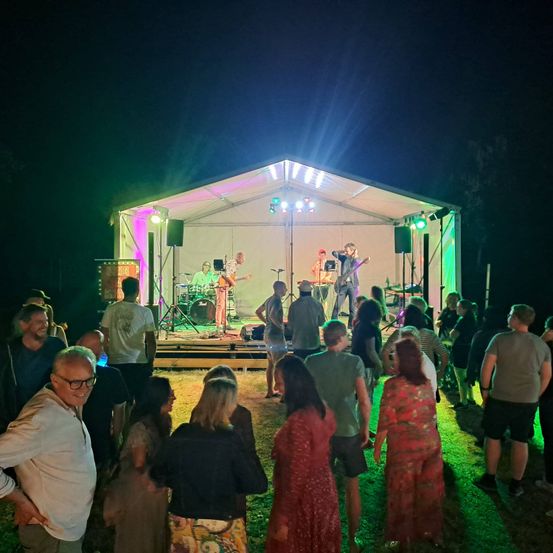 Eine Band spielt auf einer Bühne unter einem Zelt mit bunten Lichtern, während eine Menge Menschen bei Nacht zuschaut.