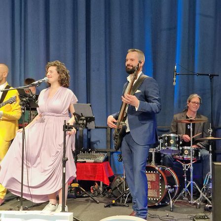 Eine Band tritt auf einer Bühne mit blauem Vorhang im Hintergrund auf. Eine Frau in einem rosa Kleid singt, während ein Mann in einem blauen Anzug die Gitarre spielt. Ein anderer Mann in einem gelben Anzug spielt ebenfalls Gitarre neben ihr. Dahinter spielt ein Schlagzeuger Trommeln und Becken.