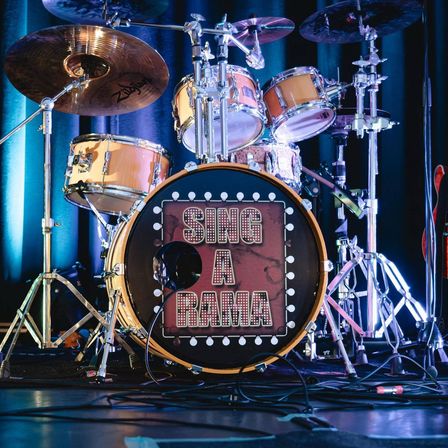 Ein Schlagzeug auf der Bühne mit Becken und einem Bass-Drum, auf dem 'SING A RAMA' in rot-weißen Buchstaben steht.