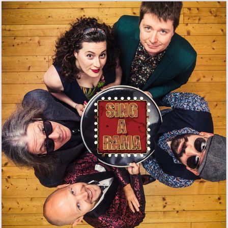 Fünf Menschen lächeln in die Kamera. Vier von ihnen halten ein Schild mit der Aufschrift 'Sing a Rama'. Der Hintergrund ist hölzern.