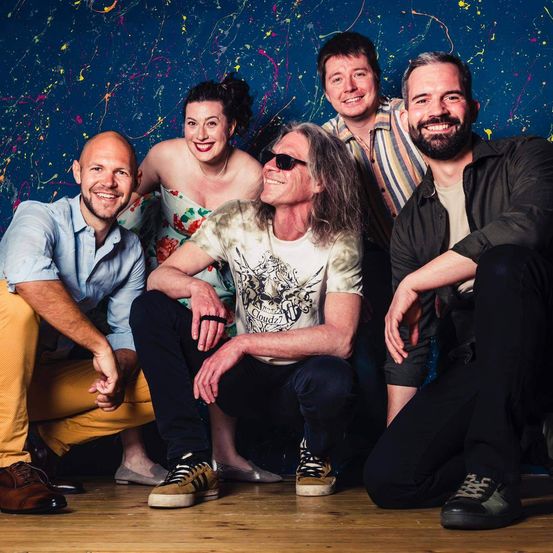 Fünf Personen posieren für ein Foto vor einem Hintergrund mit bunten Farbspritzern. Sie alle lächeln.