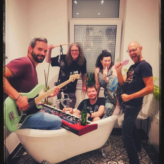 Eine Band von Musikern posiert in einer Badewanne und spielt Instrumente. Einer spielt eine grüne Gitarre, ein anderer ein Keyboard, während andere Trommelstöcke halten. Ein Mann sitzt lächelnd in der Wanne.