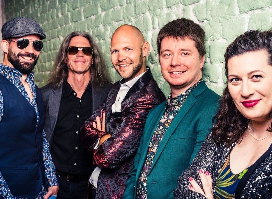 Fünf Personen, vier Männer und eine Frau, stehen eng beieinander gegen eine grüne Ziegelwand und lächeln alle. Ein Mann trägt eine Mütze und eine Sonnenbrille, während ein anderer einen kahlen Kopf hat.