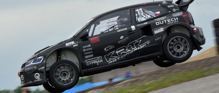 Ein schwarzer Rallye-Wagen mit der Aufschrift 11 Fucik ist in der Luft, wahrscheinlich bei einem Rennevent. Der Wagen hat Sponsorenlogos wie MTL, Berga und OPTIX. Er befindet sich über einem Grasfeld mit verschwommenen Figuren.