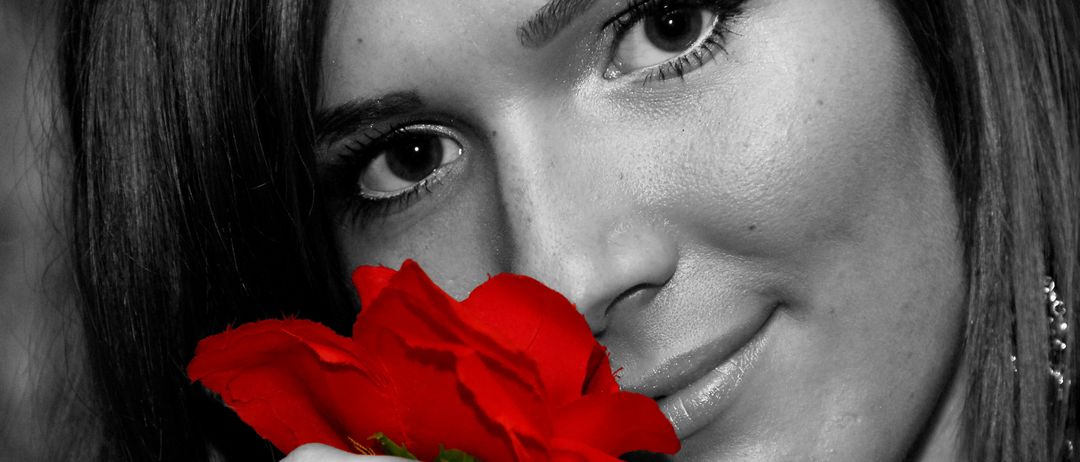 Eine Frau mit dunklem Haar hält eine rote Rose in der Nähe ihres Gesichts und lächelt. Ihre Augen sind auf die Rose gerichtet.