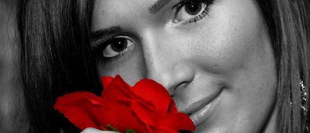 Eine Frau mit dunklem Haar hält eine rote Rose in der Nähe ihres Gesichts und lächelt. Ihre Augen sind auf die Rose gerichtet.