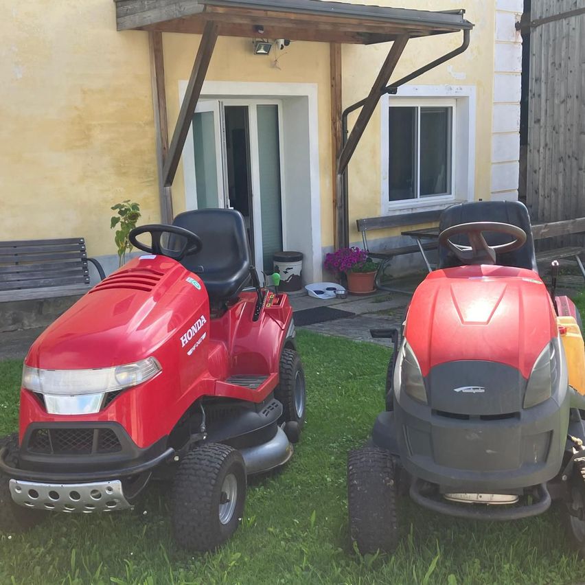 Zwei rote Rasenmäher stehen vor einem Haus mit einer Veranda, einer Tür und einem Fenster. In der Nähe befinden sich eine Bank, eine Topfpflanze und ein Mülleimer.