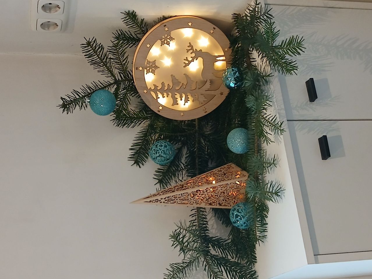 Ein Weihnachtskranz mit Tannenzweigen und einer runden Lichtinstallation mit Schneeflocken- und Rehendesign, beleuchtet mit gelben Lichtern. Blaue Zierkugeln hängen von den Zweigen.