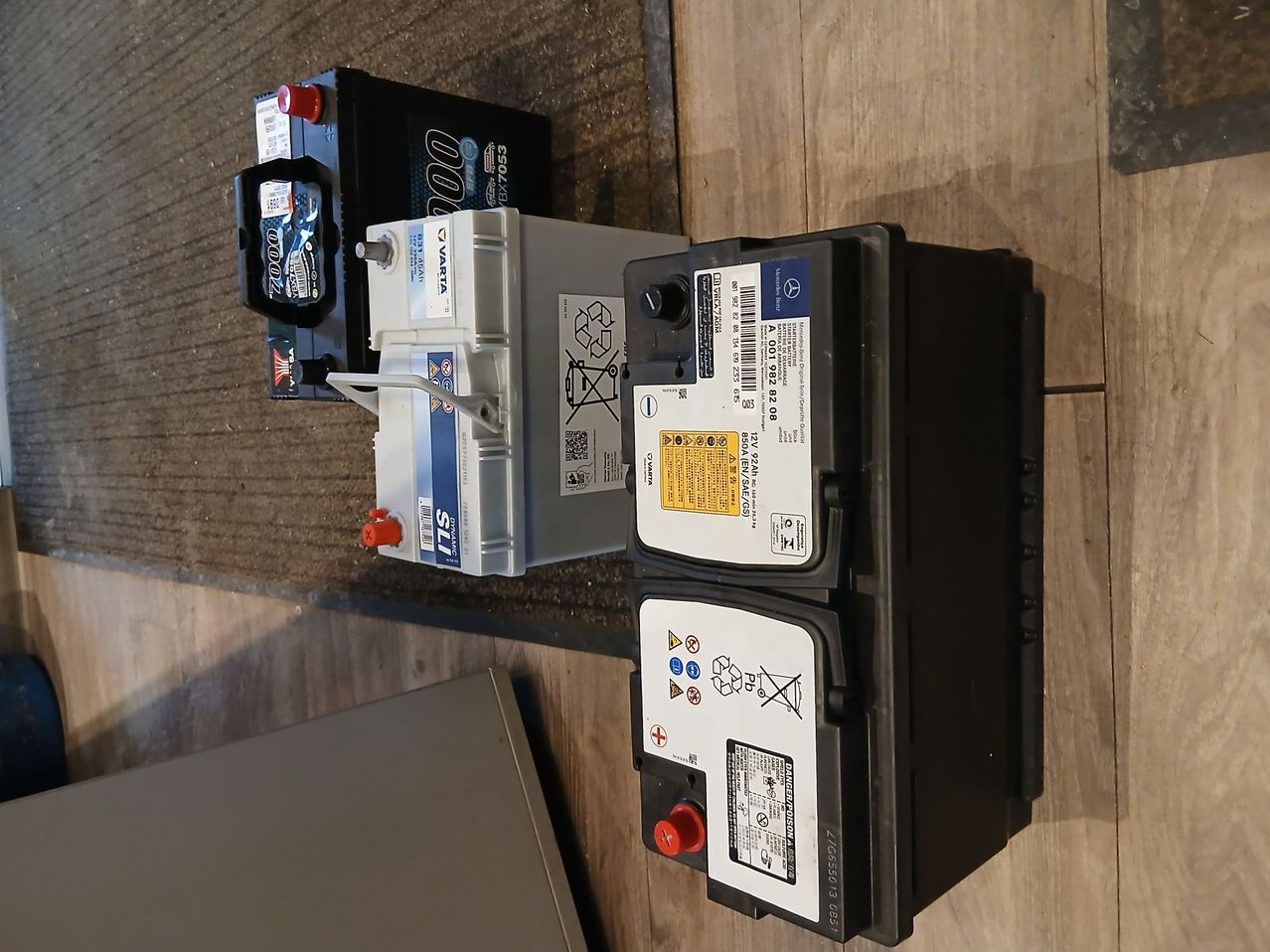 Drei Autobatterien auf einer Holzoberfläche. Zwei sind schwarz mit weißen Etiketten und roten Kappen, und eine ist weiß mit blauen Etiketten. Die weiße Batterie hat ein Recycling-Symbol.