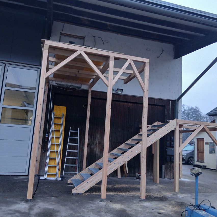 Eine hölzerne Treppe wird vor einem Gebäude errichtet. Eine gelbe Leiter und eine silberne Leiter stehen in der Nähe. Ein Auto ist vor dem Gebäude geparkt.