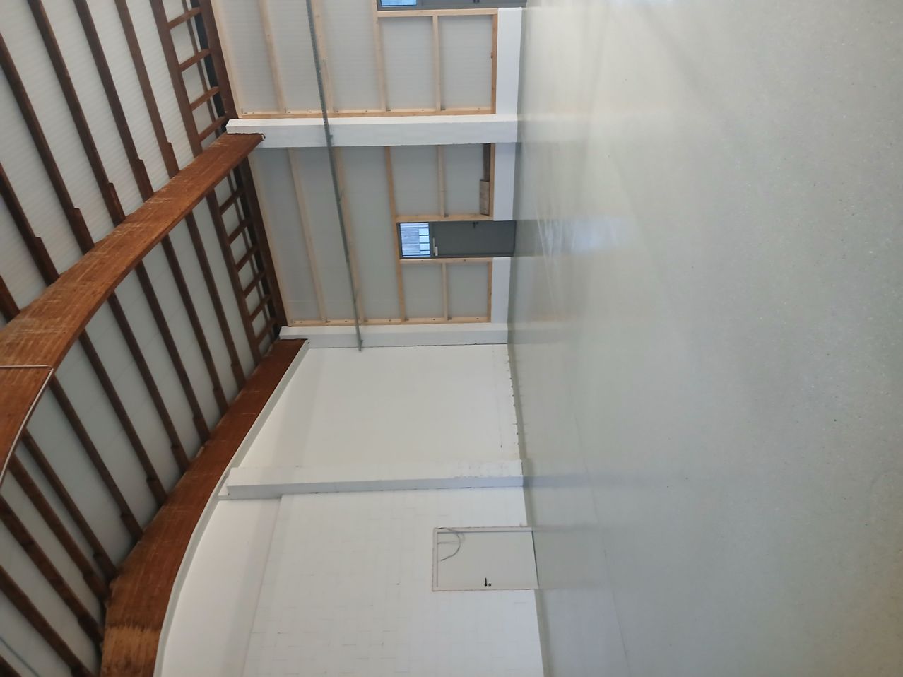 Ein leerer Raum mit einem geschwungenen hölzernen Balkon auf der linken Seite. Weiße Wände und Decke mit Holzbalken. Ein kleines Fenster befindet sich in der Mitte der Decke.