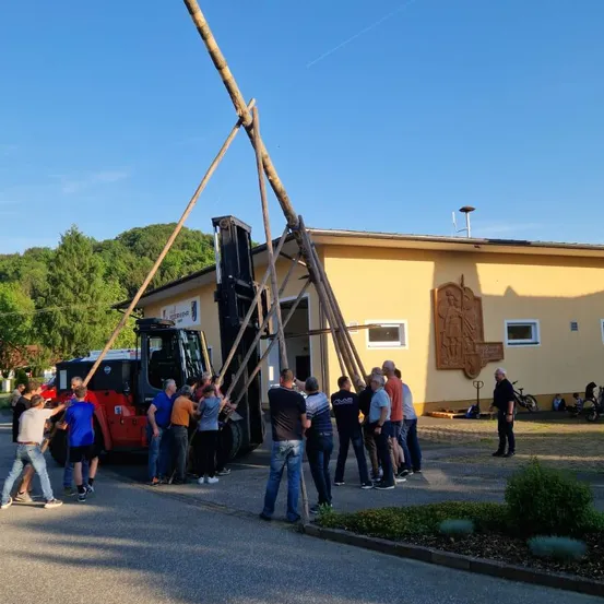 Eine Gruppe von Menschen hilft einem Gabelstapler, einen Holzpfahl mit einem großen dreieckigen Rahmen anzuheben, möglicherweise für ein Bauprojekt.