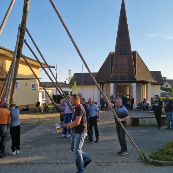 Männer in Jeans und T-Shirts versammeln sich auf einem Hof unter einem sonnigen Himmel. Ein Mann mit einem Bier stößt mit einem anderen Mann an. Eine Kirche ist im Hintergrund zu sehen.