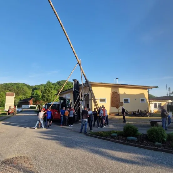 Eine Gruppe von Menschen versammelt sich um einen großen Pfahl, der von einem Fahrzeug angehoben wird. Der Pfahl wird von Holzplanken gestützt. Im Hintergrund steht ein gelbes Gebäude mit mehreren Fenstern und vor dem Gebäude geparkten Fahrrädern.