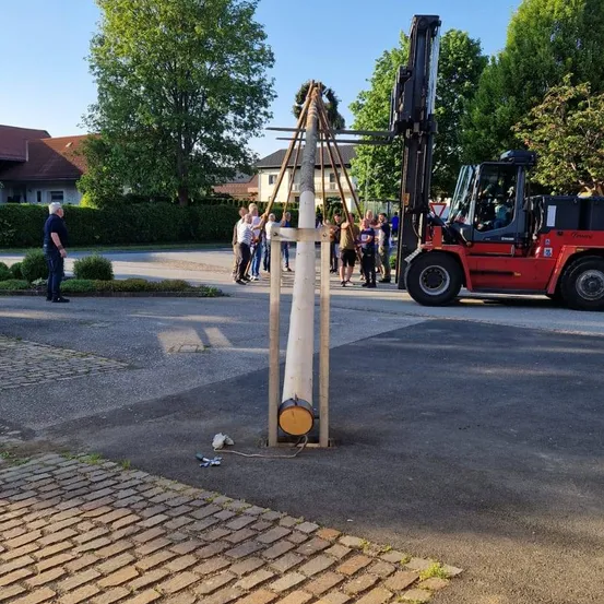 Eine Gruppe von Menschen steht um eine hölzerne Struktur mit einem großen Baumstamm auf einer Straße. Ein Gabelstapler ist neben der Struktur geparkt, der Fahrer ist drin. Bäume und Häuser sind im Hintergrund.