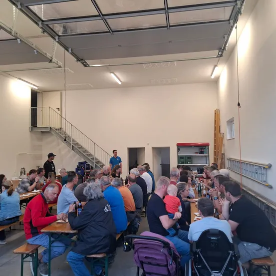 Eine große Gruppe von Menschen sitzt an Tischen in einem Speisesaal, wobei einige Personen im Hintergrund stehen. Der Raum hat weiße Wände, Metalldecken und mit weißen Tüchern bedeckte Tische. Eine Person in einem schwarzen Shirt geht auf einen Tisch mit einem Baby zu, während eine andere Person in einem blauen Shirt vor einer Treppe steht.