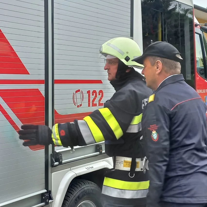 Zwei Feuerwehrleute in Uniform stehen bei einem Fahrzeug, einer trägt einen grünen Helm.