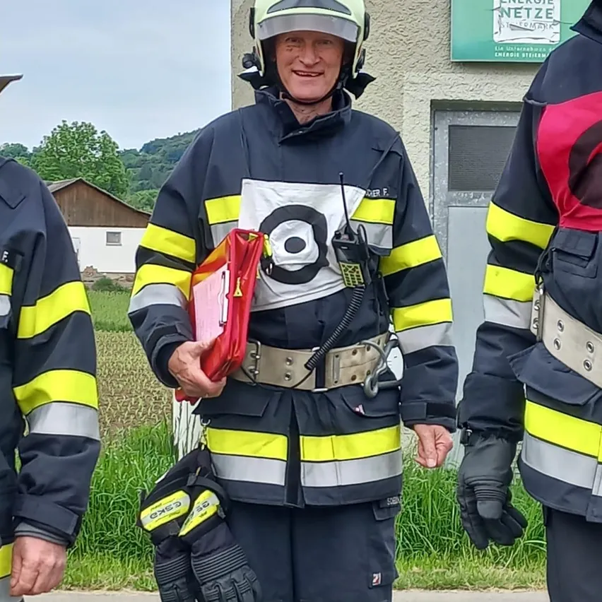 Drei Feuerwehrleute stehen vor einem Gebäude, einer hält eine rote Mappe. Sie tragen alle Helme und Uniformen.