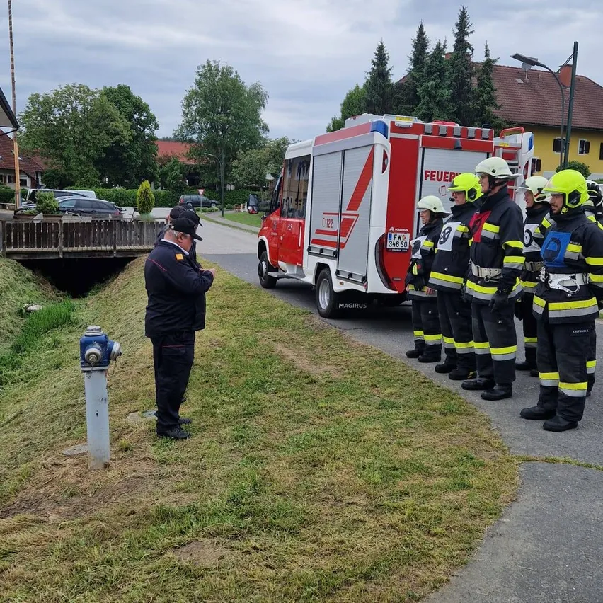 Feuerwehrleute stehen neben einem roten Feuerwehrwagen, ein Mann in Uniform inspiziert einen Feuerhydranten. Im Hintergrund sind Bäume und Häuser zu sehen.