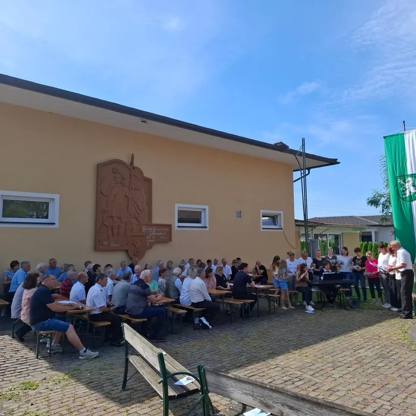 Eine Gruppe von Menschen sitzt um Tische herum vor einem Gebäude mit einem Wandbild, einer Flagge und einem klaren Himmel. Ein Chor steht vor einem Notenständer.
