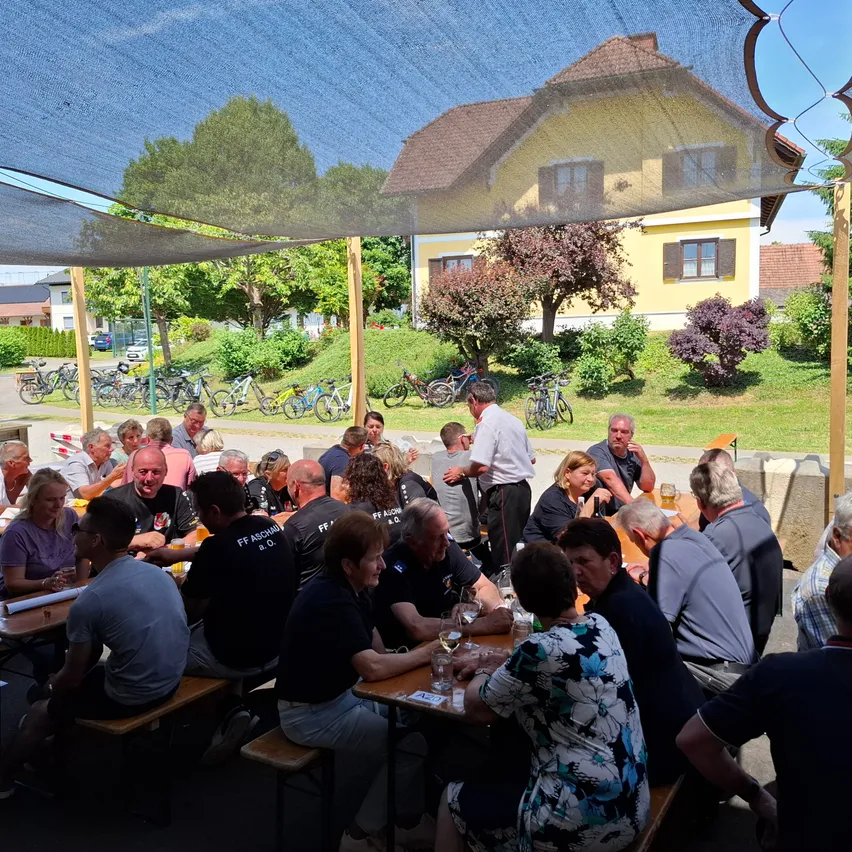 Eine Gruppe von Menschen sitzt unter einem Zelt um Tische herum draußen. Im Hintergrund befindet sich ein gelbes Haus mit geparkten Fahrrädern davor.