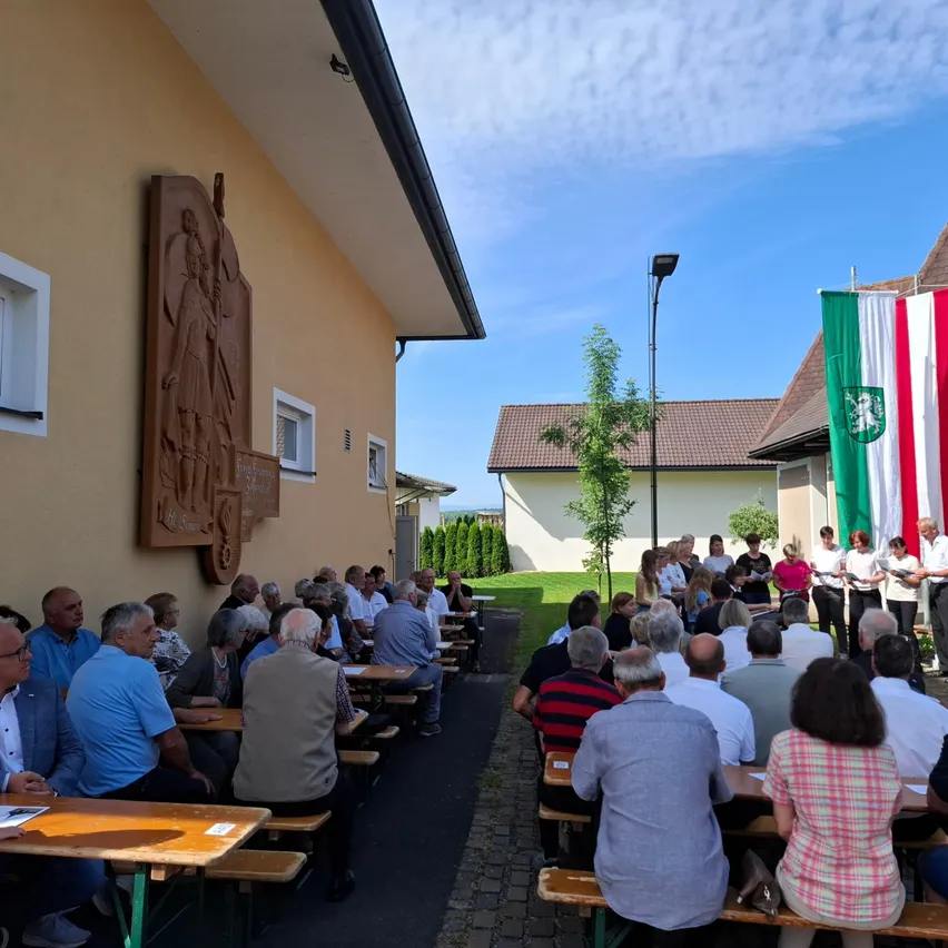 Eine Gruppe von Menschen sitzt an Tischen vor einem Gebäude unter einem klaren Himmel, mit wehender Flagge im Hintergrund.