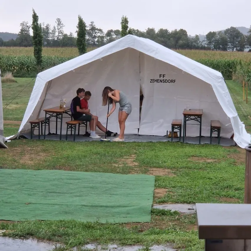 Eine Frau spielt Golf mit einem Putter in einem weißen Zelt auf einer Wiese mit zwei Männern, die an einem Tisch sitzen. Das Zelt trägt den Text 'FF ZEHENSDORF'.