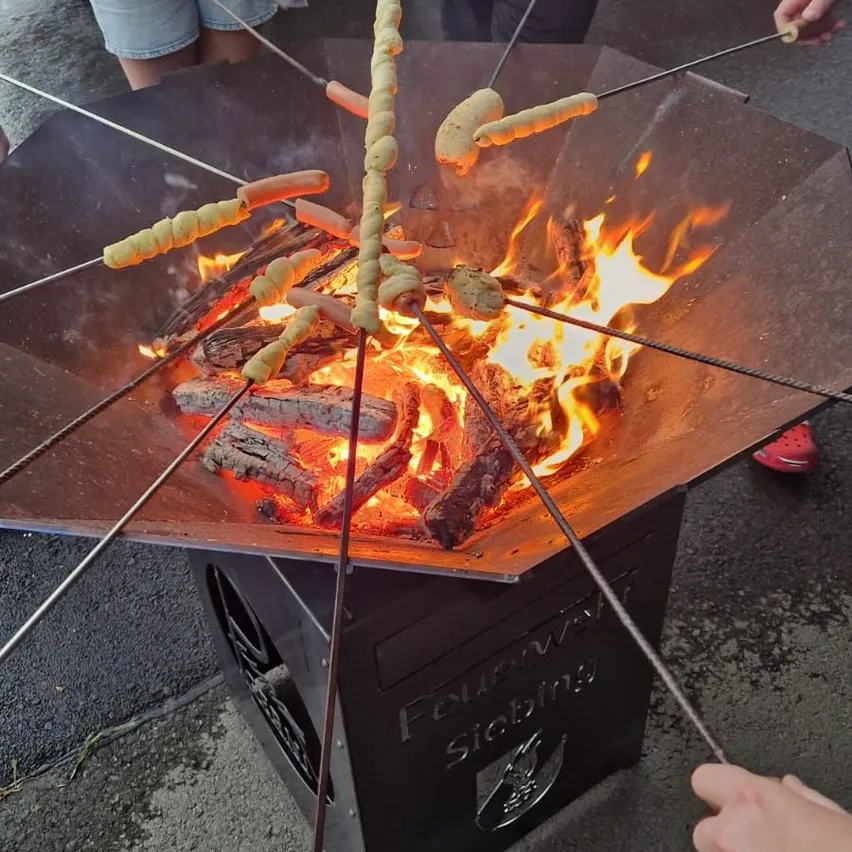 Eine Gruppe von Menschen röstet Würstchen auf Metallstäben über einem Feuer in einem achteckigen Grill im Freien. Der Grill hat einen Metalldeckel und steht auf einer schwarzen Metallbasis.