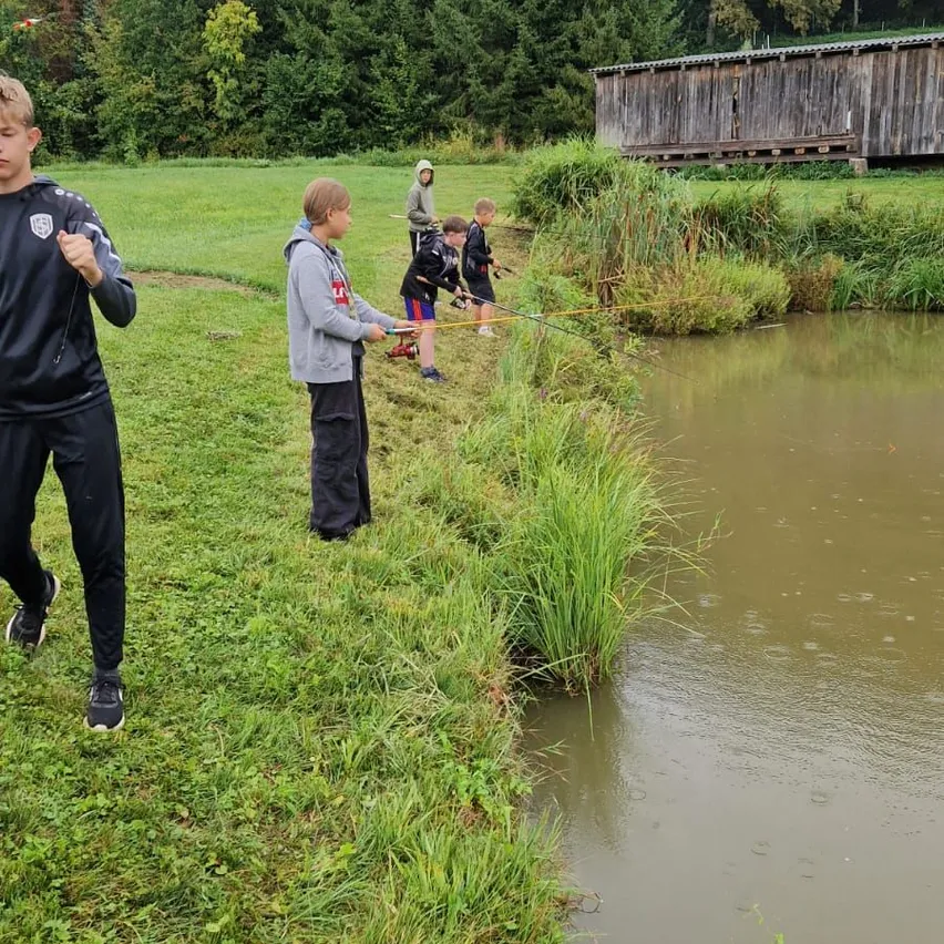 Mehrere Jungen angeln in einem Teich, mit hohem Gras am Ufer. Ein Junge in einer grauen Kapuzenjacke hält eine Angelrute. Bäume und ein Holzgebäude sind im Hintergrund.