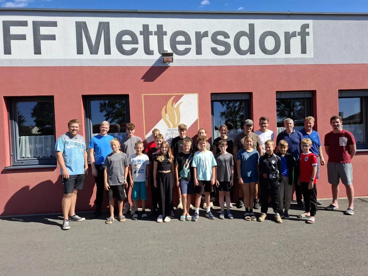 Eine Gruppe von Kindern und Erwachsenen steht vor einem Gebäude mit dem Namen 'Mettersdorf' darauf. Sie alle lächeln und scheinen für ein Foto zu posieren.
