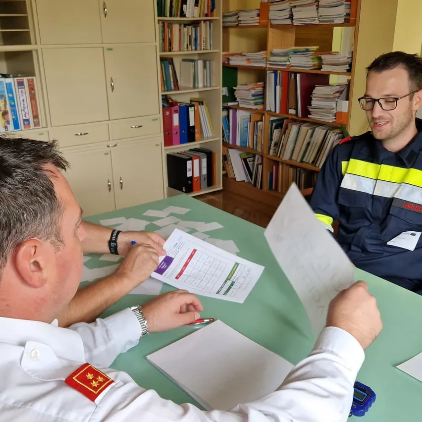 Zwei Männer in Uniformen sitzen an einem Tisch und überprüfen Papiere. Der Mann auf der linken Seite trägt einen Aufnäher mit einem Stern und einem Abzeichen. Ein Bücherregal voller Bücher steht hinter ihnen.
