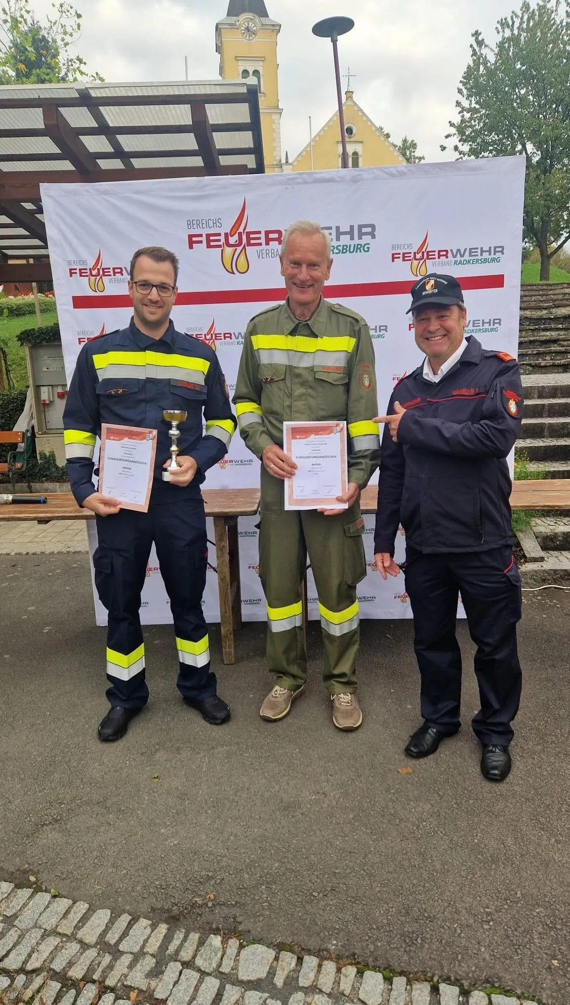 Drei Männer stehen vor einem Banner mit der Aufschrift 'Feuerwehr Verband Radkersburg'. Der Mann auf der linken Seite hält einen Pokal und ein Zertifikat. Der Mann in der Mitte hält nur ein Zertifikat. Der Mann auf der rechten Seite zeigt auf das Zertifikat des Mannes in der Mitte. Hinter ihnen steht ein Holztisch.