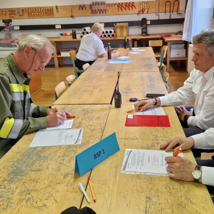 Mehrere Personen sitzen an einem langen Tisch in einem Klassenzimmer und sind mit Schreibaktivitäten beschäftigt. Ein blaues Schild mit der Aufschrift 'BSP 1' steht auf dem Tisch. Verschiedene Papiere, Stifte und eine rote Mappe befinden sich ebenfalls auf dem Tisch. Im Hintergrund sind Schreibtische mit Gegenständen und eine Ampel zu sehen.