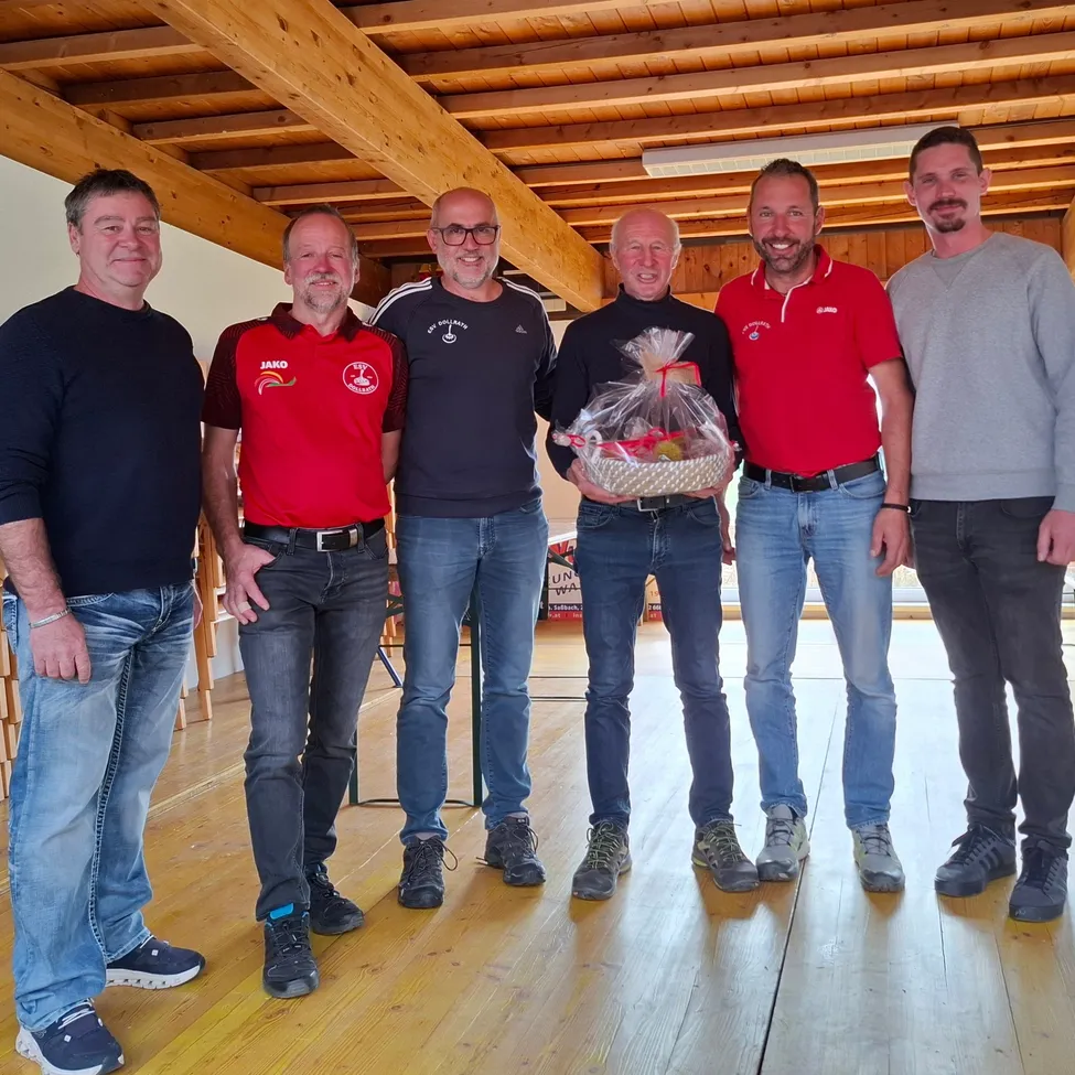 Fünf Männer stehen zusammen in einem Raum mit Holzbalken an der Decke. Der Mann in der Mitte hält einen Korb mit einem Geschenk darin.