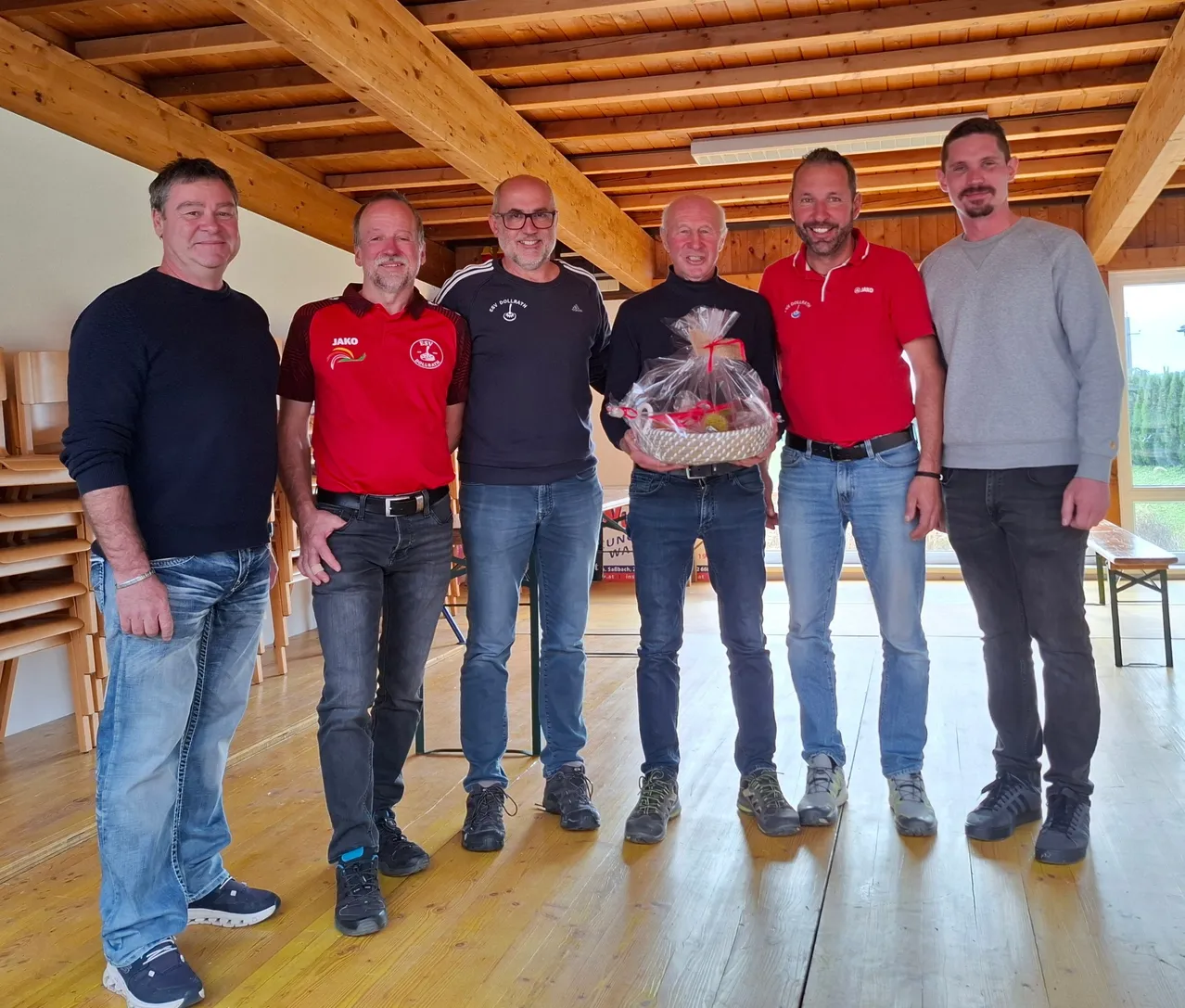 Fünf Männer stehen zusammen in einem Raum mit Holzbalken an der Decke. Der Mann in der Mitte hält einen Korb mit einem Geschenk darin.