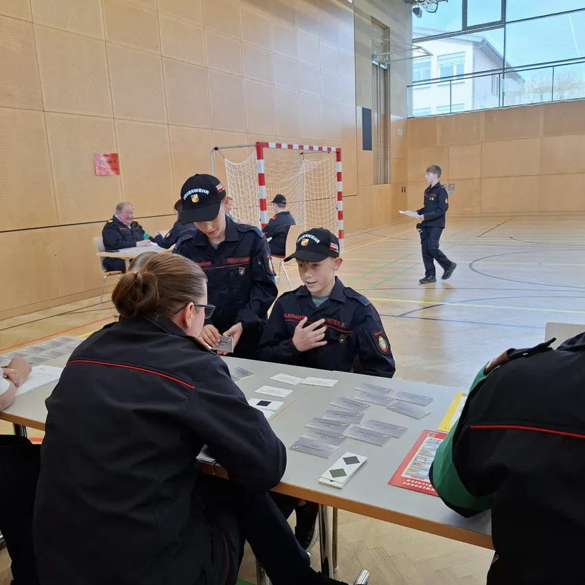 In einer Turnhalle versammeln sich mehrere Personen in Uniformen um einen Tisch und spielen Karten. Ein Junge in der Mitte spricht mit einer Frau. Hinter ihnen geht eine Person mit einem Papier.