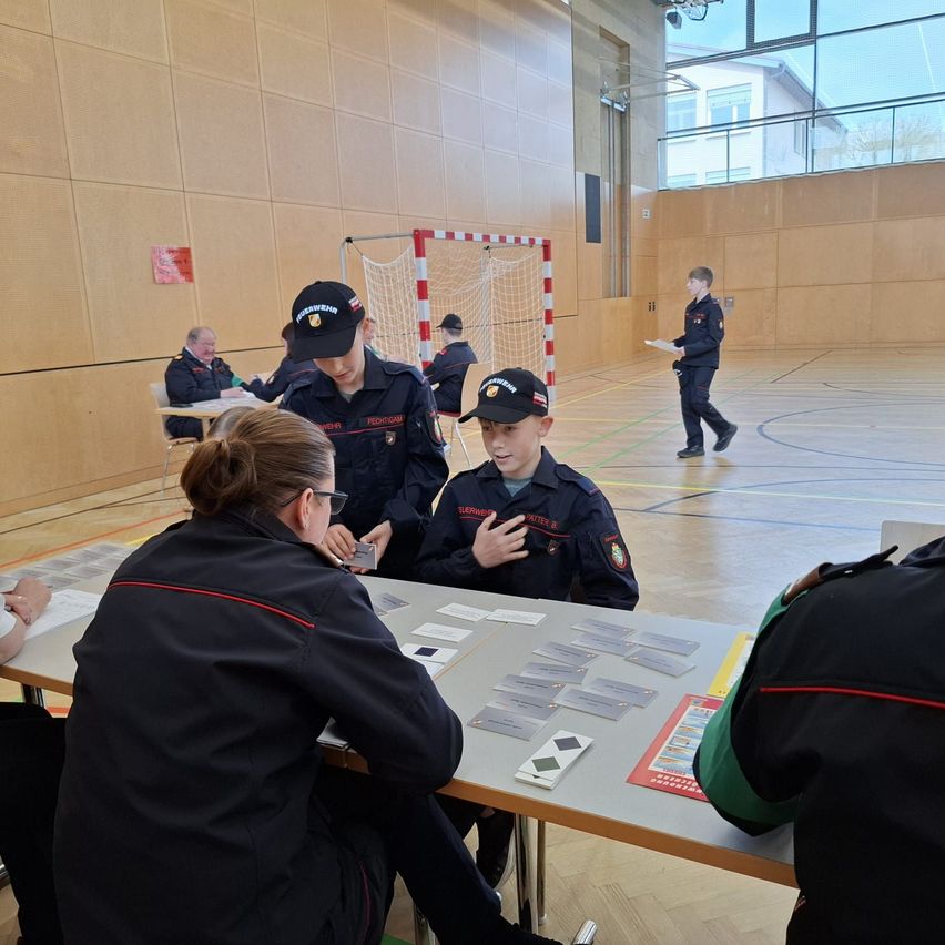 Inside a gymnasium, a group of people dressed in uniforms are gathered around a table, engaged in a card game. A boy in the center is talking to a woman. Behind them, a person walks with a paper.