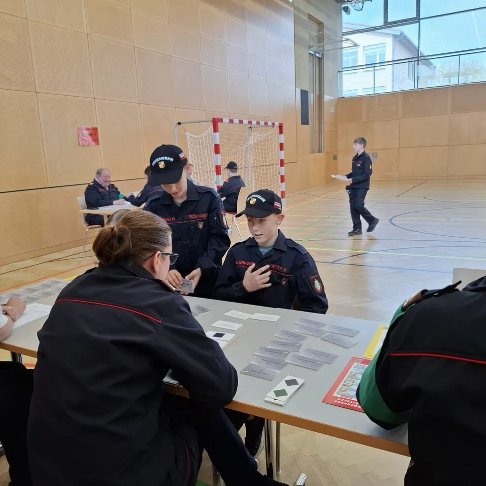 In einer Turnhalle versammeln sich mehrere Personen in Uniformen um einen Tisch und spielen Karten. Ein Junge in der Mitte spricht mit einer Frau. Hinter ihnen geht eine Person mit einem Papier.