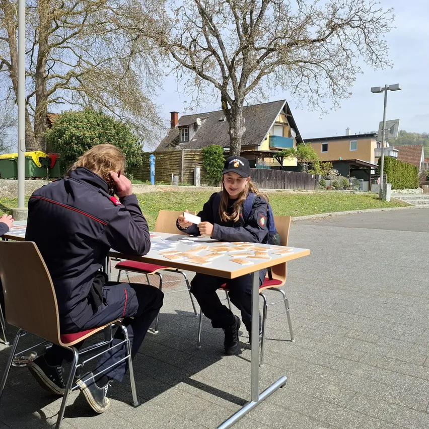 Zwei Personen sitzen an einem Tisch im Freien. Eine hält eine Karte, die andere scheint am Telefon zu sprechen. Hinter ihnen sind Häuser und Bäume zu sehen.