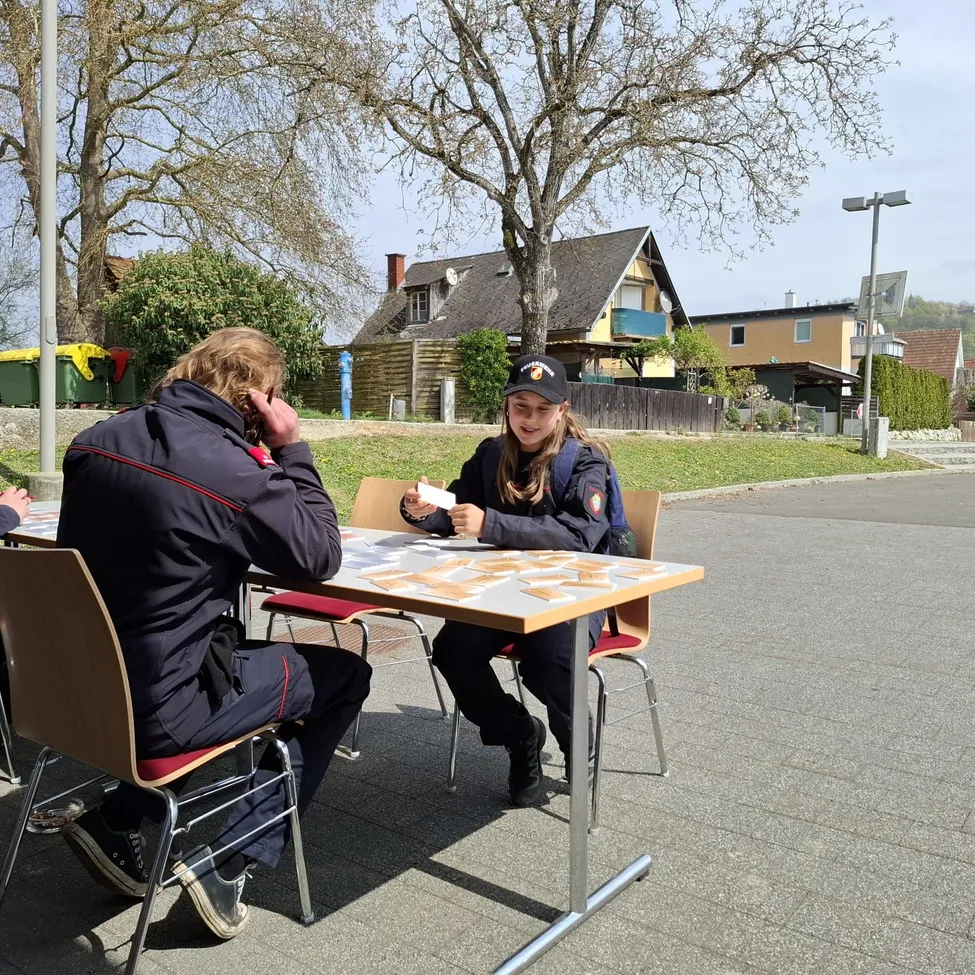 Zwei Personen sitzen an einem Tisch im Freien. Eine hält eine Karte, die andere scheint am Telefon zu sprechen. Hinter ihnen sind Häuser und Bäume zu sehen.