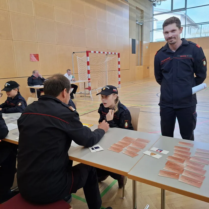 Eine Gruppe von Personen in Uniformen sitzt an Tischen mit Karten. Eine Frau spricht mit einem Mann. Ein anderer Mann steht und hält ein Papier. Ein Fußballtor im Hintergrund.