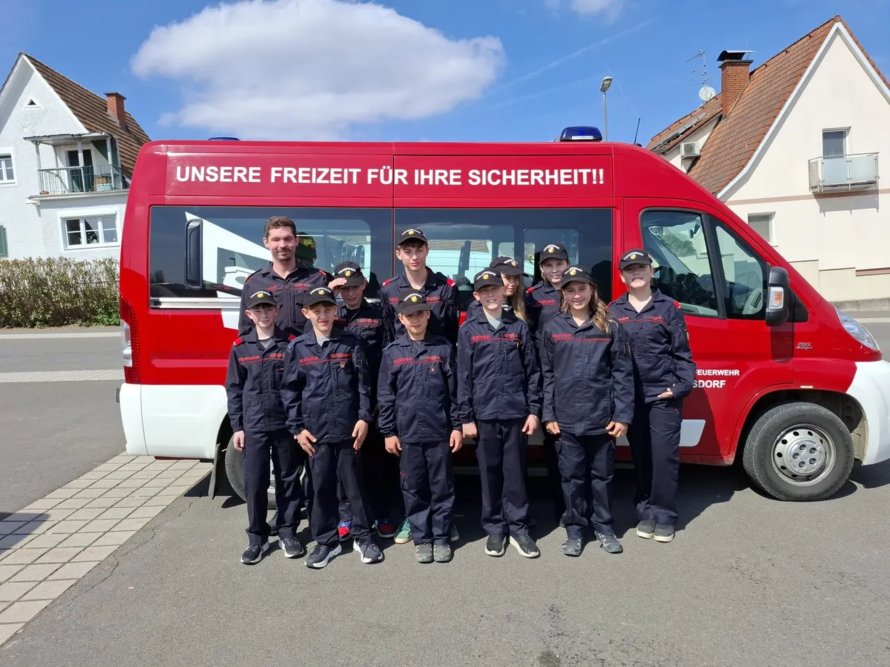 Eine Gruppe junger Menschen in marineblauen Uniformen steht vor einem roten Feuerwehrauto mit der Aufschrift 'Unsere Freiheit für Ihre Sicherheit.'