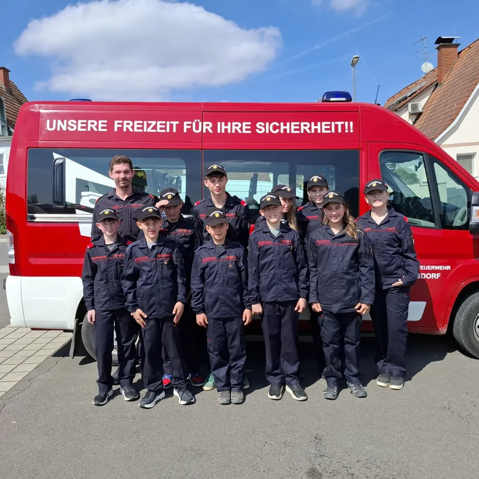 Eine Gruppe junger Menschen in marineblauen Uniformen steht vor einem roten Feuerwehrauto mit der Aufschrift 'Unsere Freiheit für Ihre Sicherheit.'