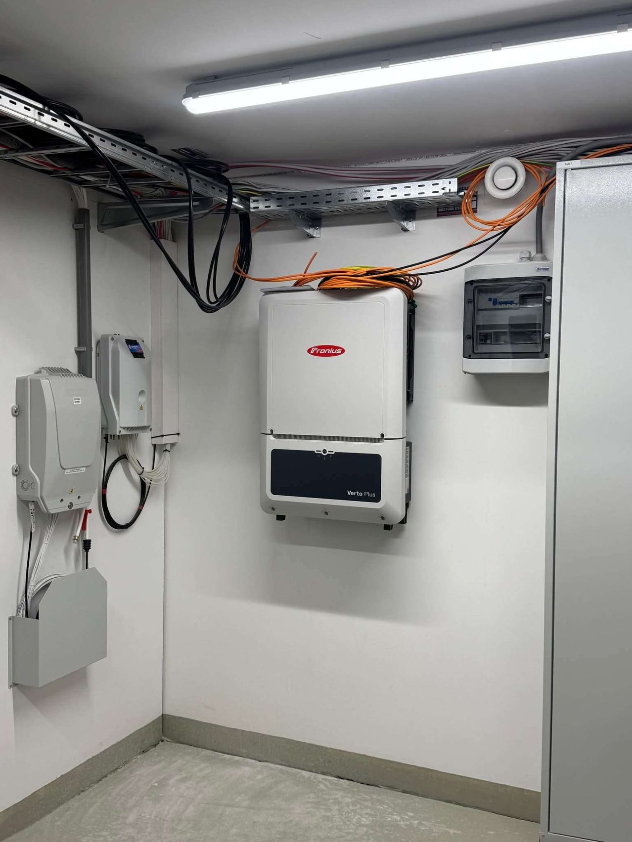 Ein Raum mit weißen Wänden enthält einen weißen Elektrokasten, einen grauen Kasten mit Kabeln und einen weißen Kasten mit einem roten Logo, der als Verto Plus gekennzeichnet ist.