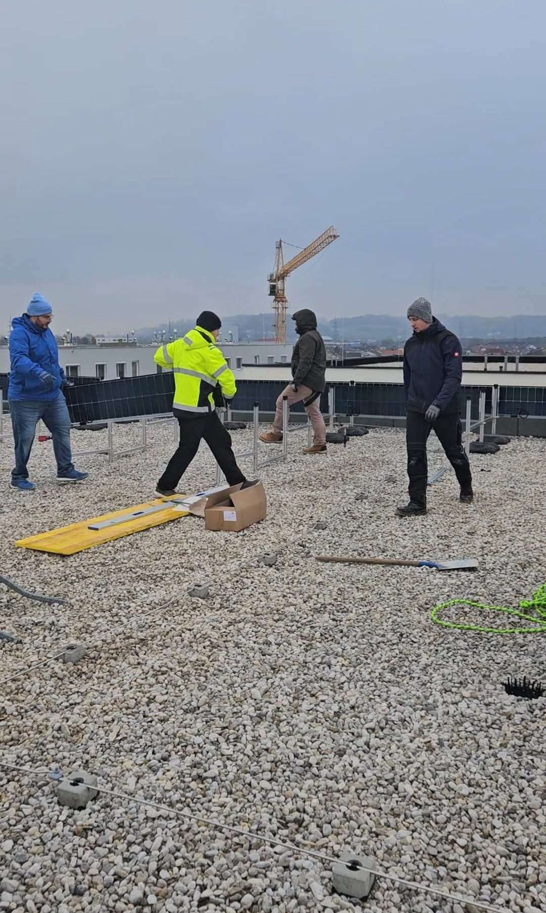 Fünf Männer in Winterkleidung arbeiten auf einem Dach. Ein Mann platziert eine gelbe Tafel. Hinter ihnen werden Solarpanele installiert. In der Ferne ist ein Kran zu sehen.
